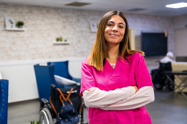 Photograph of a social care worker in bright pink scrubs standing with arms crossed in a medical or care facility. Background includes wheelchairs, blue chairs, and a person seated, indicating a patient care environment.