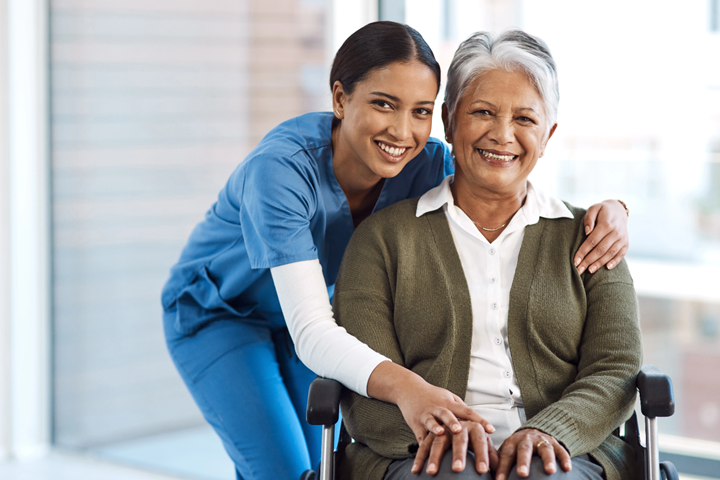 Photograph showing a healthcare worker in blue scrubs comforting an elderly person seated in a wheelchair. The setting appears to be a bright, modern medical or care facility, emphasizing support and care for elderly or disabled individuals.