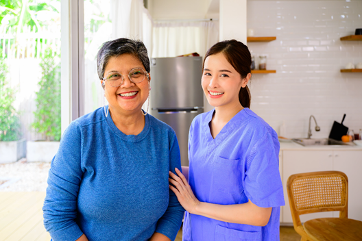 Photograph showing a healthcare worker in blue scrubs standing beside an elderly woman wearing a blue sweater in a bright kitchen setting. The healthcare worker has a supportive hand on the elderly woman's shoulder, highlighting a caregiving or medical assistance context.