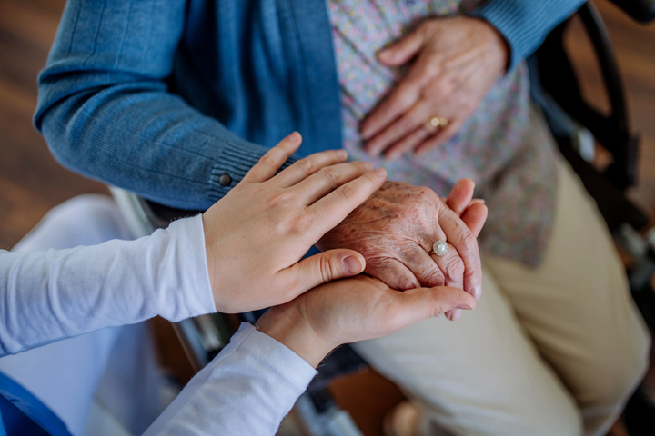 Photograph showing a younger person gently holding and comforting an elderly person's hand, who is seated and wearing a blue cardigan and floral dress. The image conveys care and support, highlighting the contrast between smooth and wrinkled skin and the presence of a ring on the elderly person's finger.