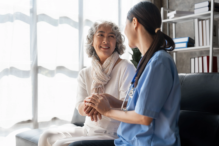 Photograph showing a healthcare professional in blue scrubs holding hands and talking with an elderly woman wearing a white sweater and scarf in a bright room. The scene conveys care and support, with a background featuring shelves with books and files.